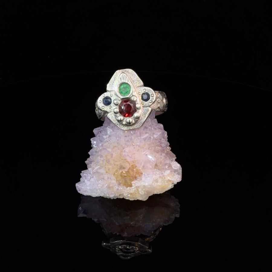 Handcrafted sterling silver size 9 ring featuring natural garnet and emerald center stones, two Montana sapphire accents, engraved face, and floral patterned band with surreal botanical design.