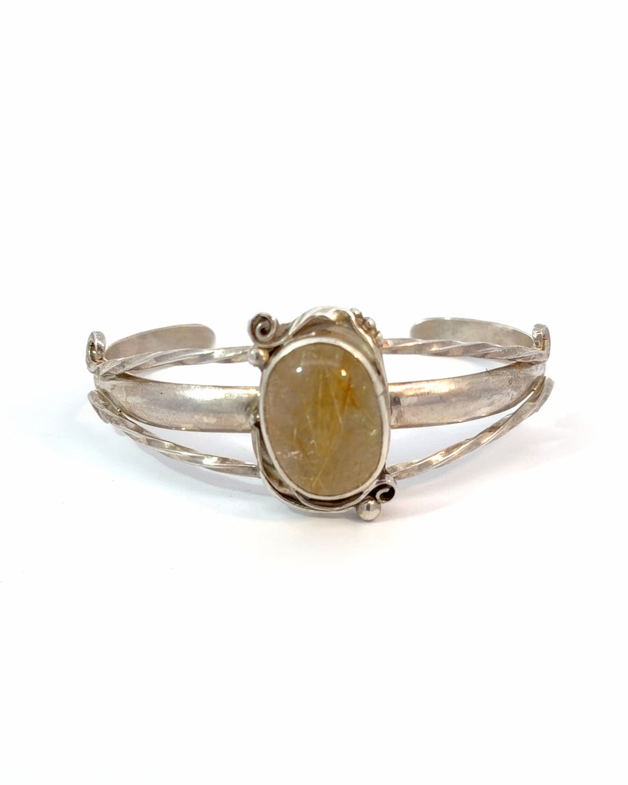 Handcrafted sterling silver bangle bracelet featuring a natural oval golden rutilated quartz cabochon with needle-like rutile inclusions, bezel setting, scrollwork, and twisted wire details.