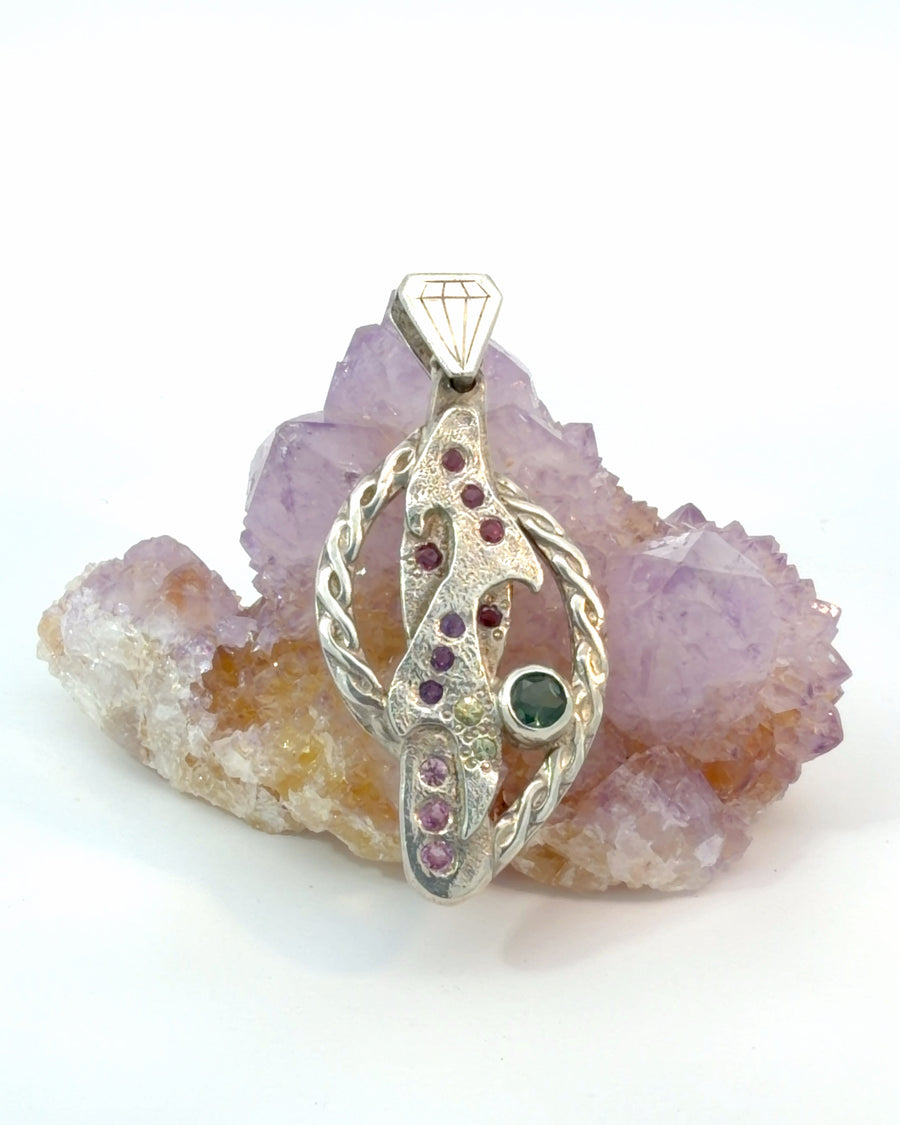 Handcrafted silver pendant featuring a twisted abstract frame with amethyst and ruby accents, bezel-set green gemstone, and engraved geometric bail.