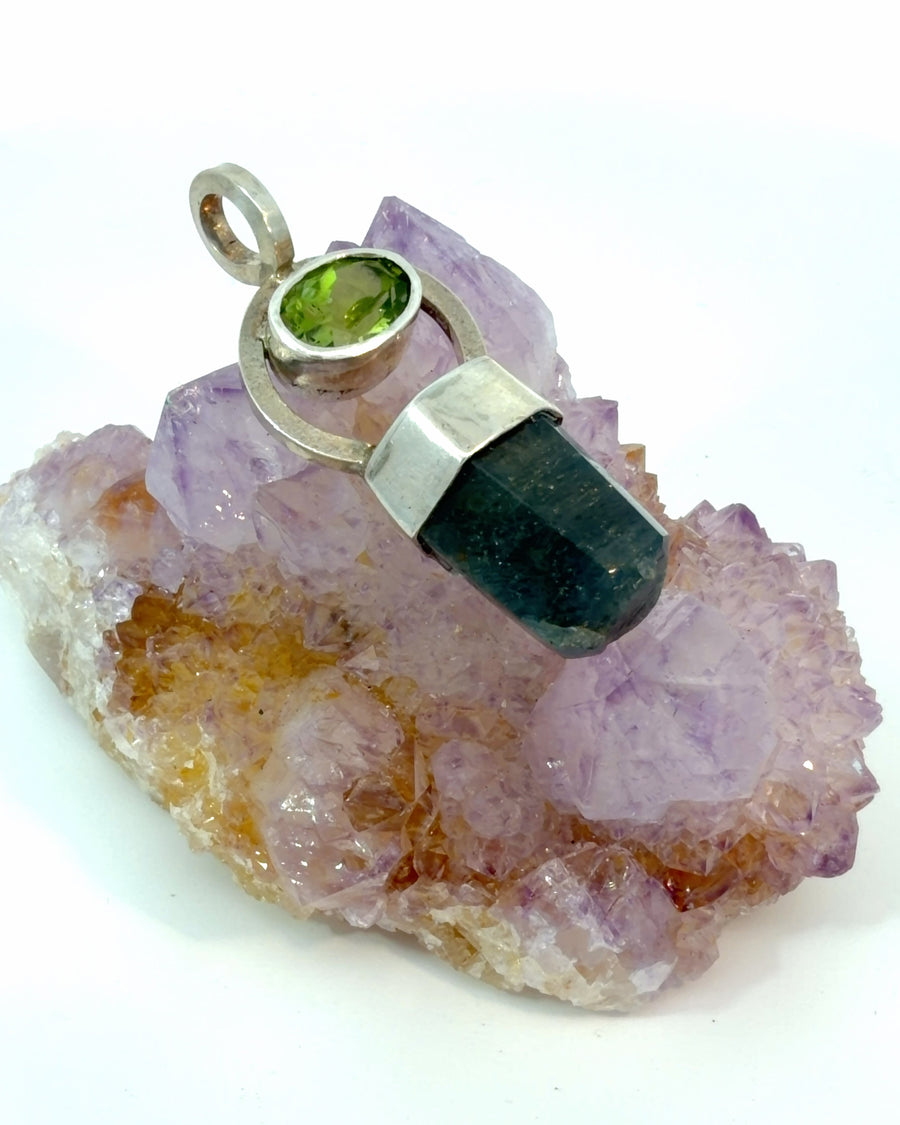 Handcrafted silver pendant featuring a bright green oval peridot in a circular frame with a natural dark blue sapphire crystal drop.
