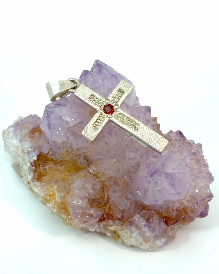 Textured silver cross pendant featuring a round deep red garnet gemstone centerpiece and polished silver bail.