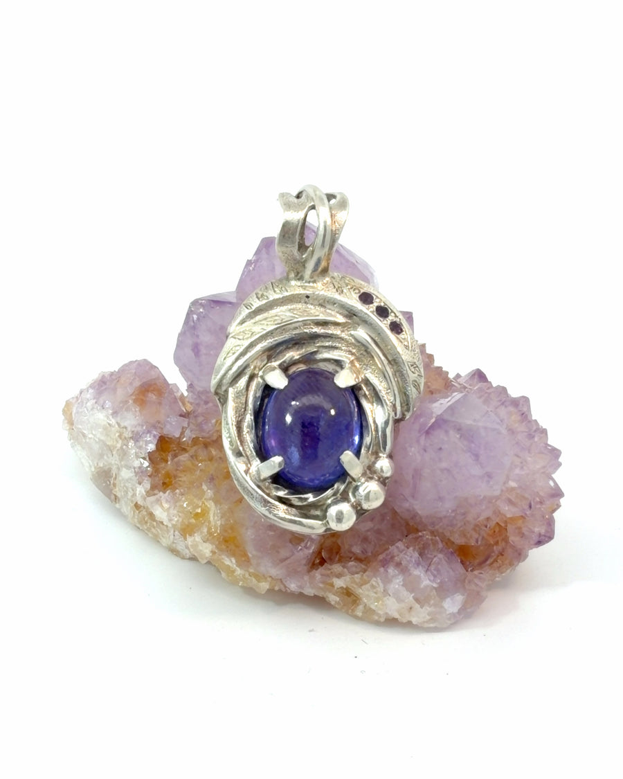 Handcrafted sterling silver pendant featuring a deep purple oval Tanzanite cabochon accented by dark Amethyst gemstones in an organic swirling silver design.