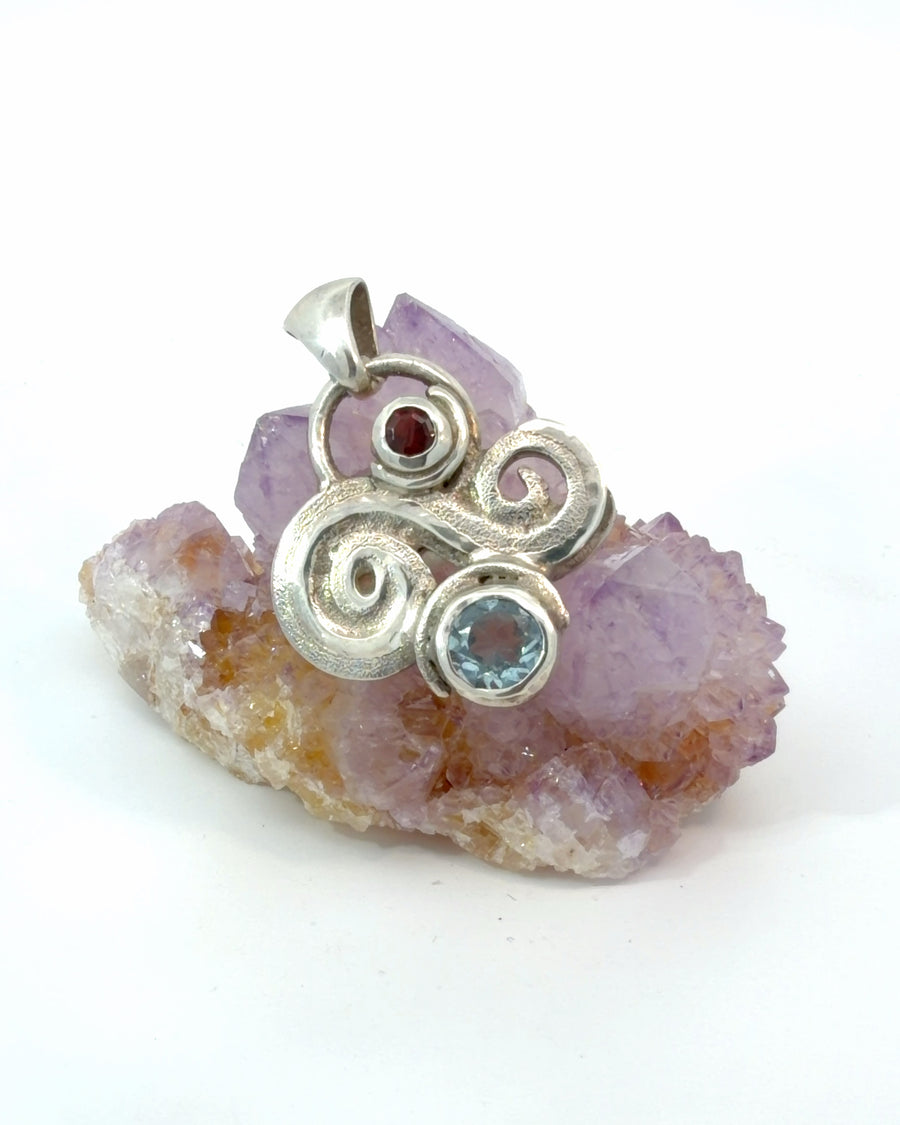 Handcrafted sterling silver swirl pendant featuring a round blue topaz gemstone and deep red garnet accent.