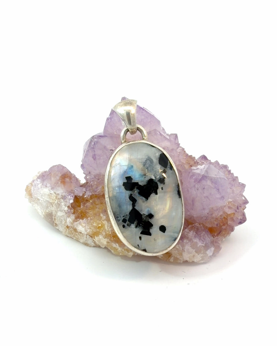 Natural oval moonstone pendant with black inclusions in a sterling silver bezel setting featuring a luminous iridescent glow.