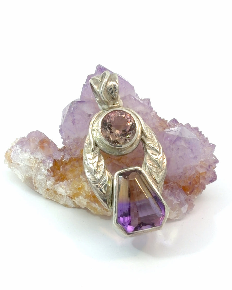 Handcrafted sterling silver botanical leaf pendant featuring a geometric faceted bi-color amethyst suspended beneath a round bezel-set pink gemstone with textured silver leaves.
Shopify Tags
