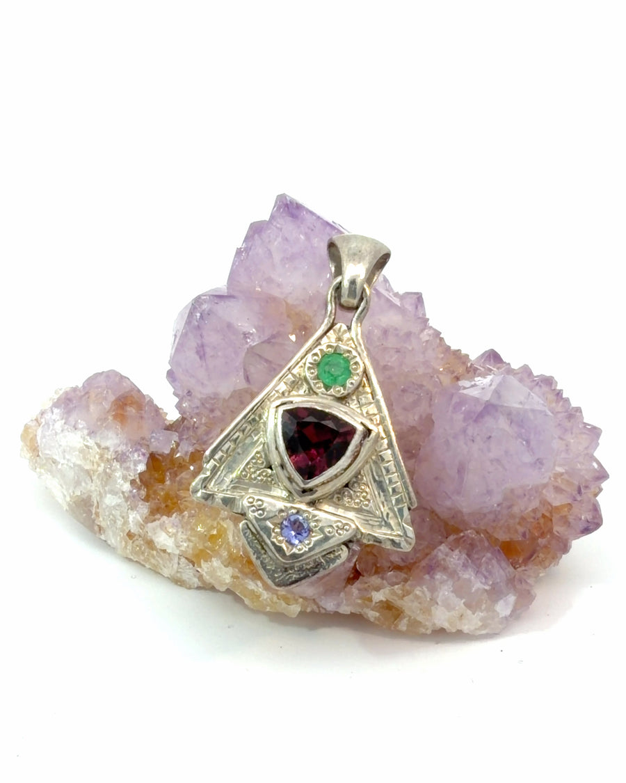 Aztec-inspired sterling silver pendant featuring a trillion-cut garnet with emerald and tanzanite accent gemstones in a geometric handcrafted design.